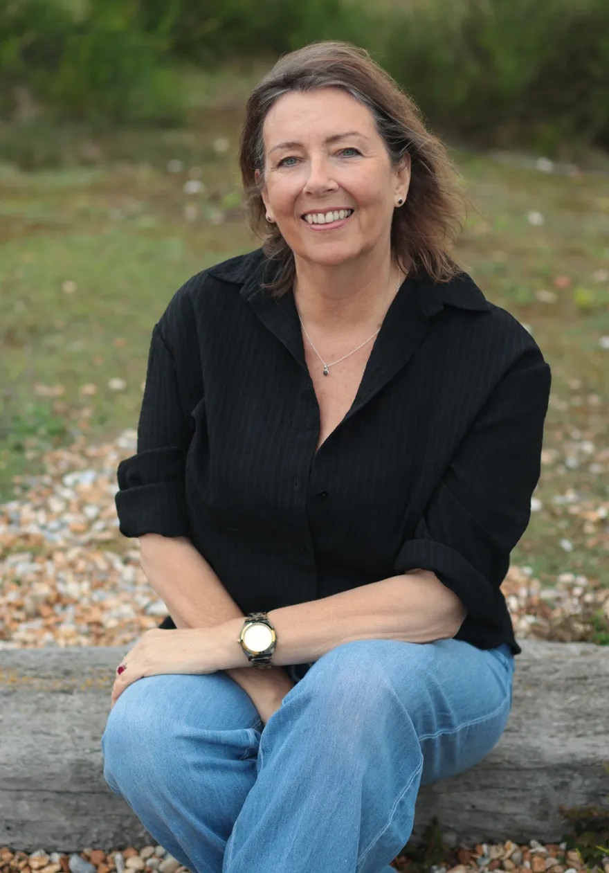 Author Lucie Bowins, smiling, seated outdoors on a shingle beach.